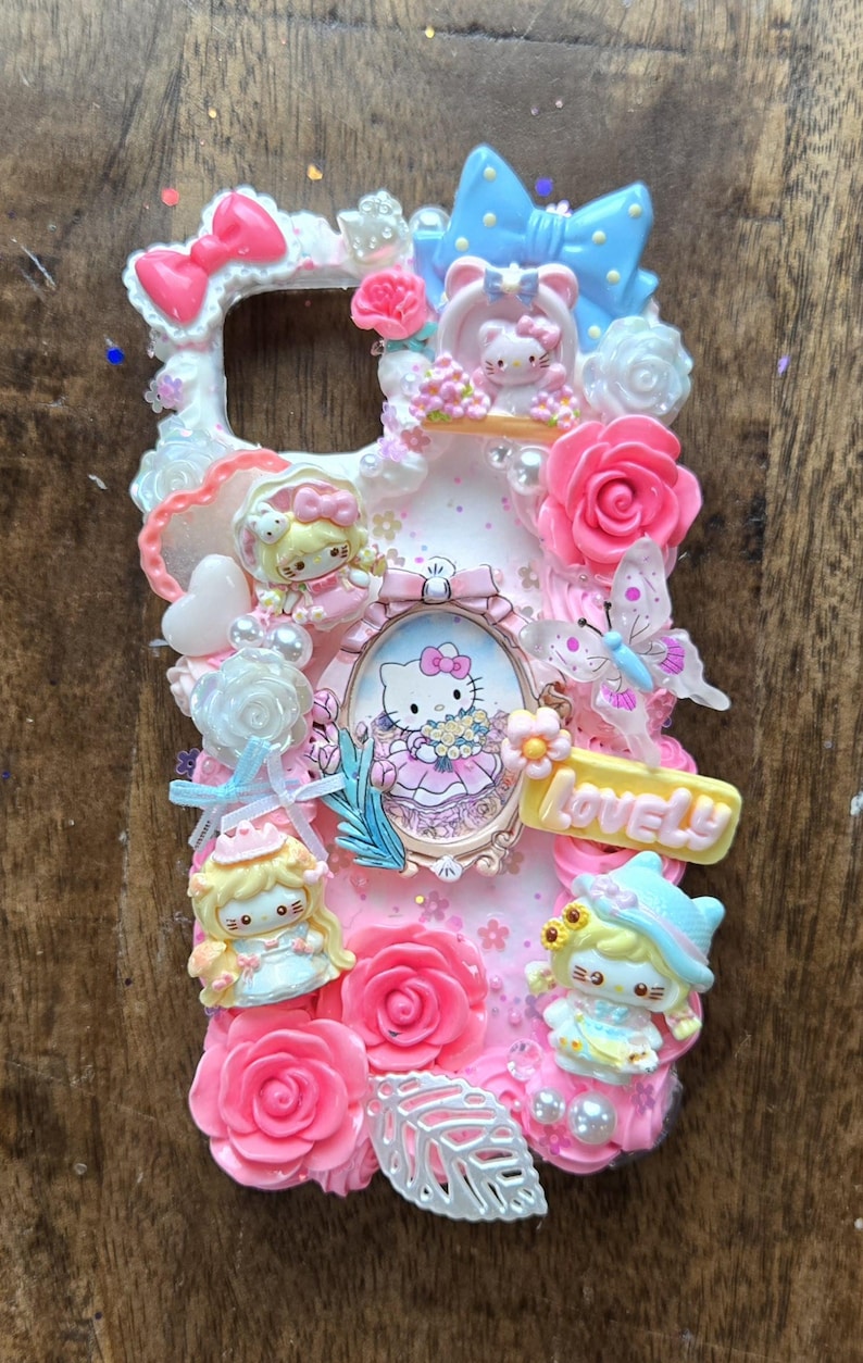 May include: A pink phone case decorated with Hello Kitty and other cartoon characters, pink roses, bows, and the word "LOVELY". The case has a white border and is covered in glitter.