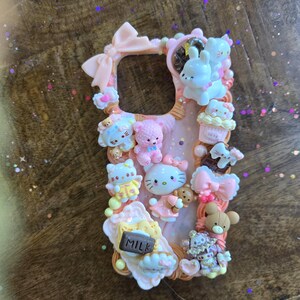 May include: Pink phone case decorated with various kawaii-style figurines, including Hello Kitty, teddy bears, and cupcakes. The case features a bow at the top and a milk carton charm. The overall design is pastel and whimsical.