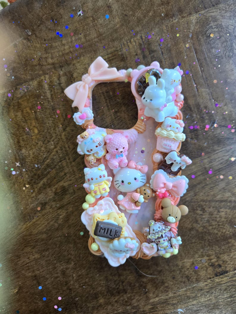 May include: Pink phone case decorated with various kawaii-style figurines, including Hello Kitty, teddy bears, and cupcakes. The case features a bow at the top and a milk carton charm. The overall design is pastel and whimsical.