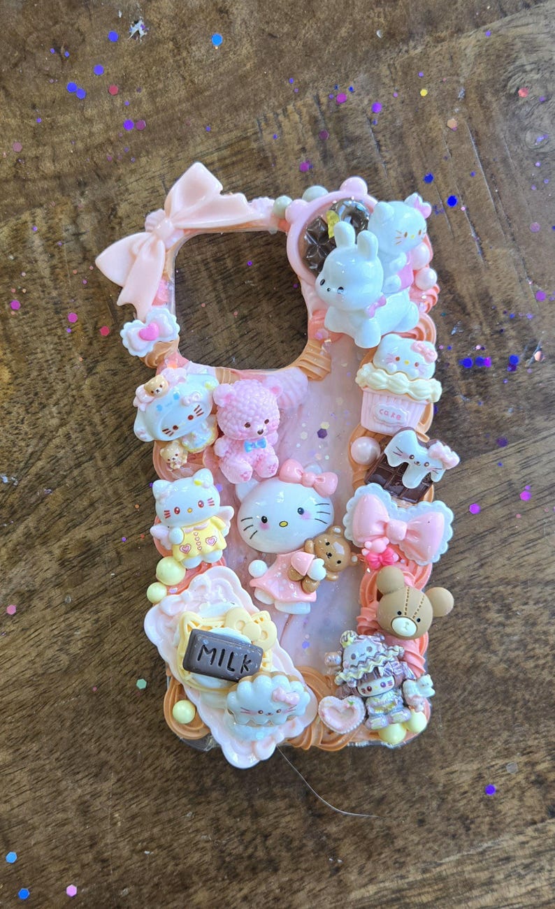 May include: A pink phone case adorned with numerous decorative elements. The case features a variety of small, colorful figurines, including Hello Kitty, teddy bears, cupcakes, and bows. The word "MILK" is visible on a small sign. The overall design is cute and whimsical.