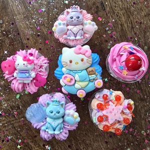 Può includere: Una collezione di cabochon decorativi in resina colorata. I cabochon presentano vari design, tra cui un gatto con una corona, un personaggio di Hello Kitty e un cupcake con una ciliegia. I cabochon sono posizionati su una superficie glitterata.