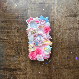 May include: A decorative phone case featuring a pink and white design with Hello Kitty characters, roses, and bows. The case has a 3D effect with various embellishments and the word "Lovely".