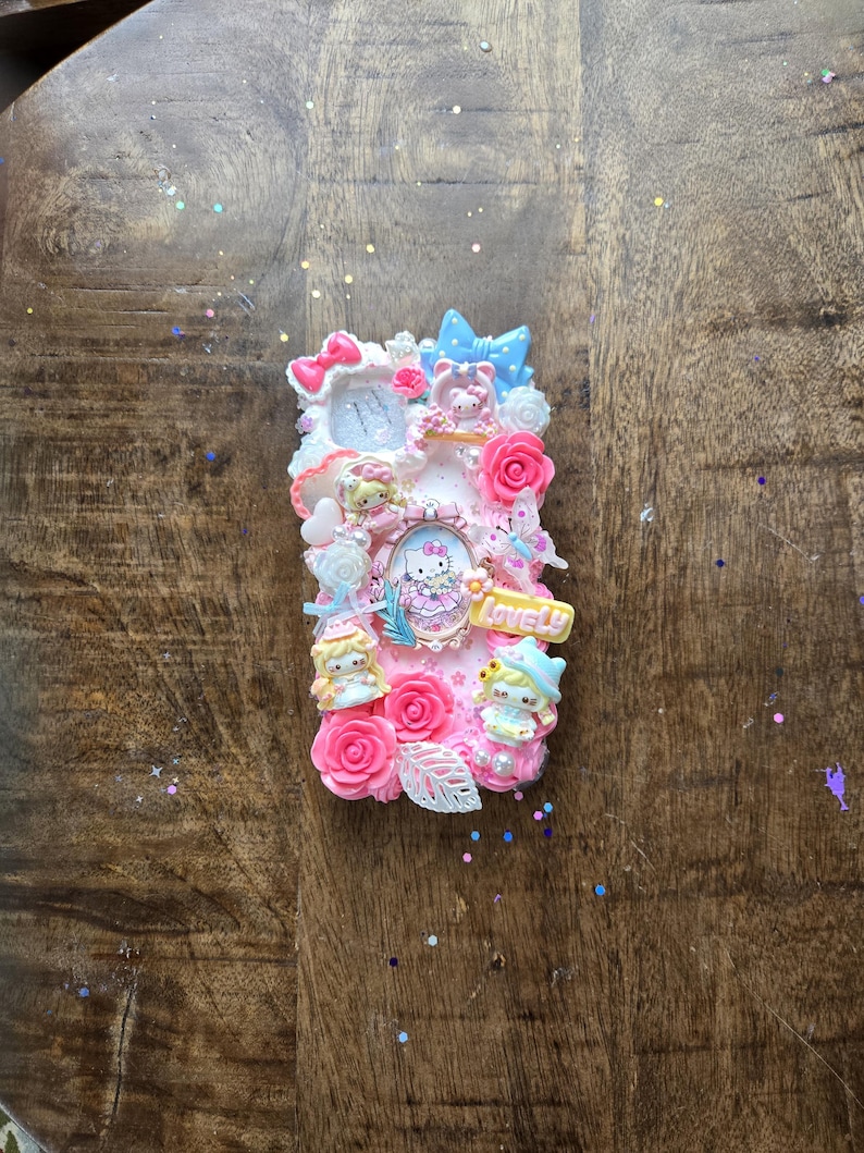 May include: A decorative phone case featuring a pink and white design with Hello Kitty characters, roses, and bows. The case has a 3D effect with various embellishments and the word "Lovely".