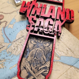 May include: A red and black wooden pendant with the words "VINLAND SAGA" in large, distressed lettering. The pendant features a detailed illustration of a character with crossed swords. The pendant is placed on a map.