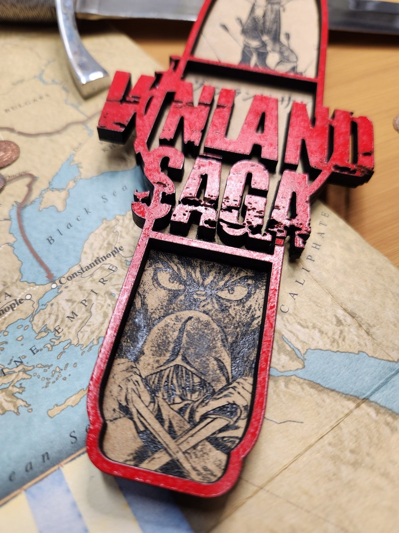 May include: A red and black wooden pendant with the words "VINLAND SAGA" in large, distressed lettering. The pendant features a detailed illustration of a character with crossed swords. The pendant is placed on a map.