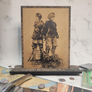 May include: A framed print featuring two figures in armor, holding swords, with the text "FINAL FANTASY TACTICS" at the base. The artwork is in black and brown tones, set against a light brown background. The frame is dark brown.