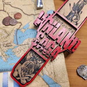 May include: A red wooden decorative piece with the text "VINLAND SAGA" in a stylized font. The piece is placed over a map with illustrations and coins. The map shows parts of Europe and Asia.