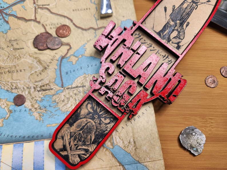 May include: A red wooden decorative piece with the text "VINLAND SAGA" in a stylized font. The piece is placed over a map with illustrations and coins. The map shows parts of Europe and Asia.