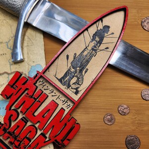 May include: A wooden decorative piece with the text "VINLAND SAGA" in red, alongside a Japanese text. The piece features a black and white illustration of a person with arrows. A sword and coins are also present.