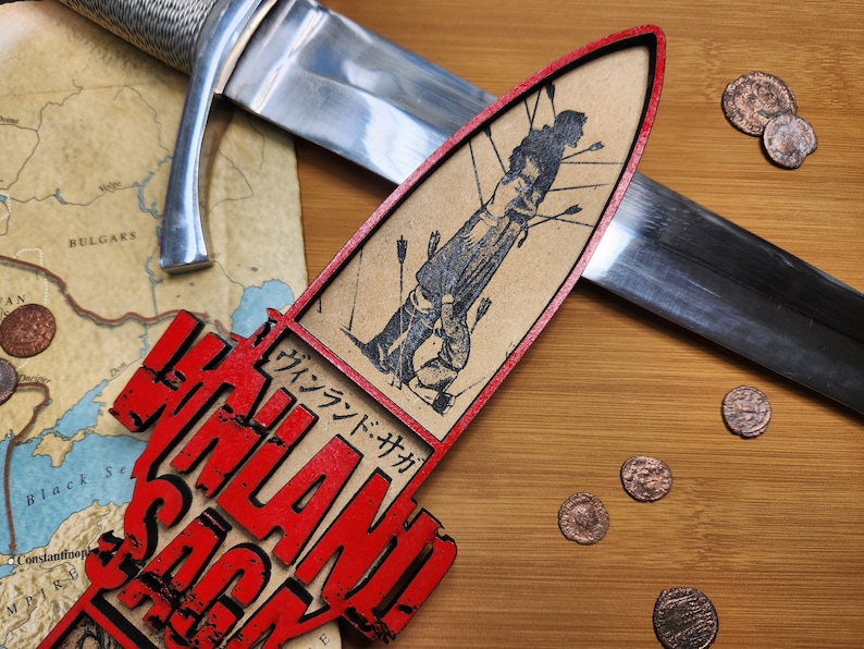 May include: A wooden decorative piece with the text "VINLAND SAGA" in red, alongside a Japanese text. The piece features a black and white illustration of a person with arrows. A sword and coins are also present.