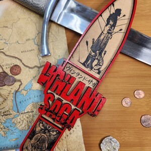May include: A decorative wooden dagger featuring the text "VINLAND SAGA" in red, with a black and white illustration of a figure pierced by arrows. A silver sword and a map are in the background.