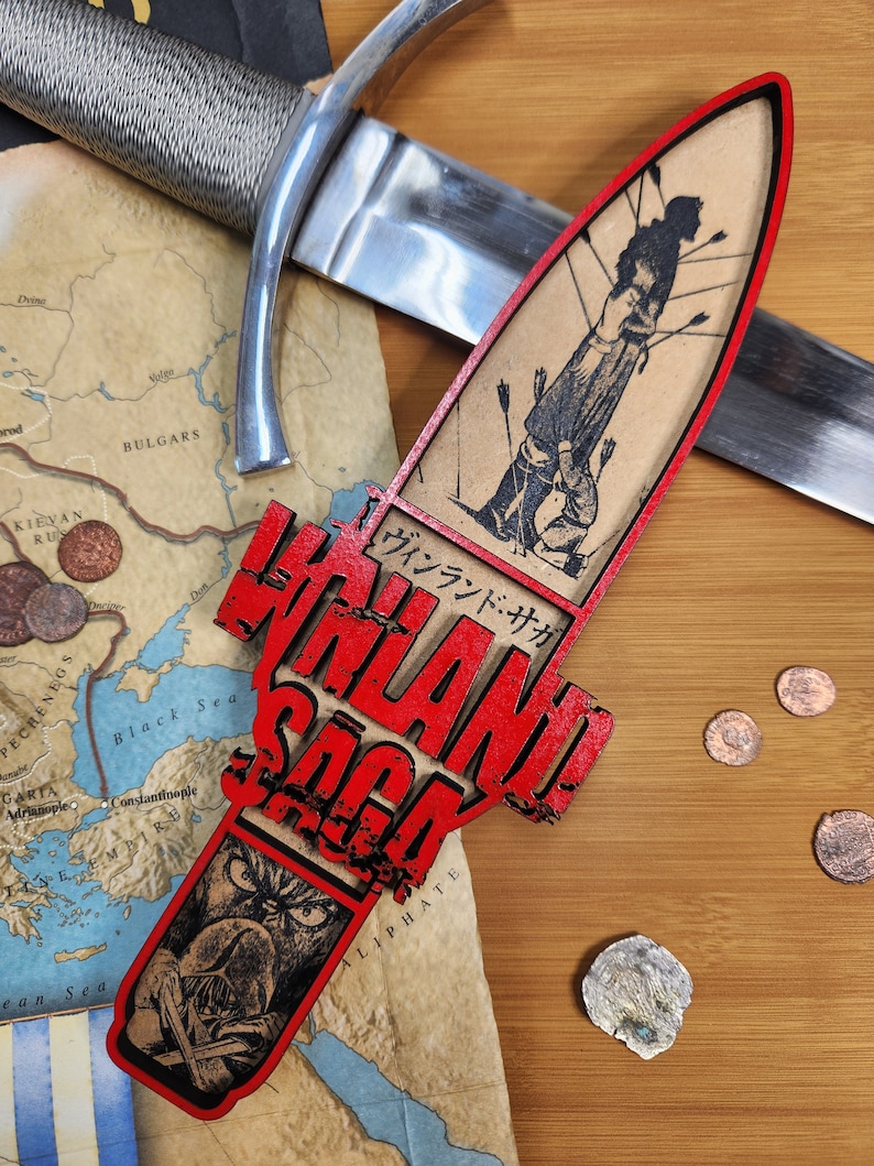 May include: A decorative wooden dagger featuring the text "VINLAND SAGA" in red, with a black and white illustration of a figure pierced by arrows. A silver sword and a map are in the background.