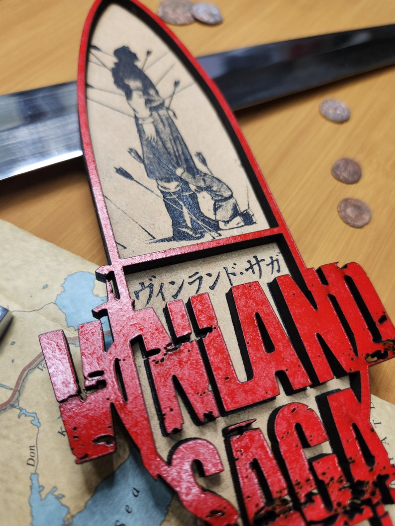 May include: A red and black wooden decorative piece with the words "VINLAND SAGA" in a distressed font. The piece features a black and white illustration of a person with arrows. Japanese text is also present.