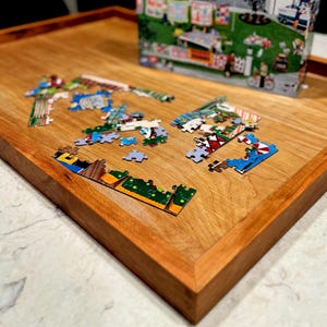 May include: A rectangular wooden puzzle board with a raised edge, featuring a smooth light brown wood grain surface. Several colorful cardboard jigsaw puzzle pieces are scattered across the center, with the original puzzle box standing upright.