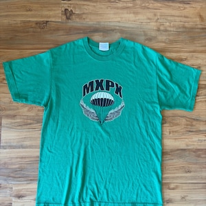 Może przedstawiać: Zielona koszulka z napisem "MXPX" nad wzorem spadochronu ze skrzydłami. Koszulka z krótkim rękawem jest wykonana z miękkiego materiału. Koszulka leży płasko na drewnianej powierzchni. Grafika jest czarno-biała.