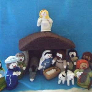 Könnte beinhalten: Eine gestrickte Weihnachtskrippe mit einem braunen Holzstall, einem gelben Engel und Figuren von Maria, Josef und dem Jesuskind. Es gibt auch Schafe und Hirten in der Szene.