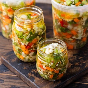 May include: Glass jars and plastic containers filled with a colorful mixture of chopped vegetables in a clear liquid. The vegetables include orange carrots, green peppers, and white cauliflower. The jars sit on a wooden cutting board.