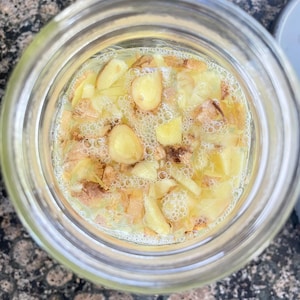 May include: A glass jar containing a yellow liquid with pieces of ginger. The ginger is in various forms, including slices and chunks. The liquid has bubbles, indicating fermentation. The jar is transparent, allowing the contents to be seen clearly.