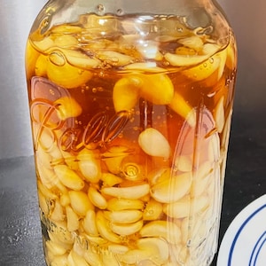 May include: A glass jar containing peeled garlic cloves in a golden liquid. The jar has a grey lid and is labelled with the word "Ball". The contents are visible through the glass. The jar is on a dark surface.