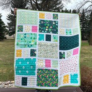 May include: A patchwork quilt with a green, white, and pink color scheme. The quilt is made up of various squares and rectangles of different fabrics, each with a unique pattern. The quilt is being held up by a person's hands.