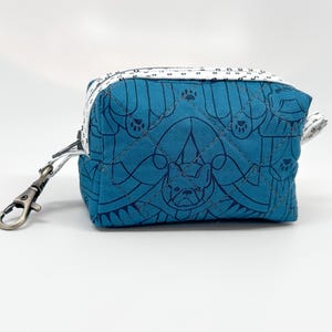 May include: A small, square teal pouch with a black dog and paw print design. The pouch has a white zipper and a silver-colored clasp. The design is quilted, and the pouch is likely made of fabric.