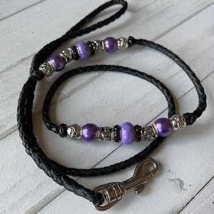 May include: A black leather dog leash with a silver clasp and purple beads. The leash is braided and has a decorative design.