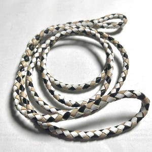 May include: Braided leather cord in a circular shape. The cord is woven with alternating sections of white, black, and tan leather. The cord is likely used for jewelry making or crafting.