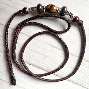 May include: Braided dark brown leather necklace with decorative beads. The necklace features a central tiger's eye bead, flanked by silver-toned and dark brown beads. The leather cord is tightly woven.