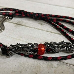 May include: Black and red braided leather leash with a silver-colored clasp. The leash features two silver dragon heads with a red bead in the center. The leash is designed for pets.