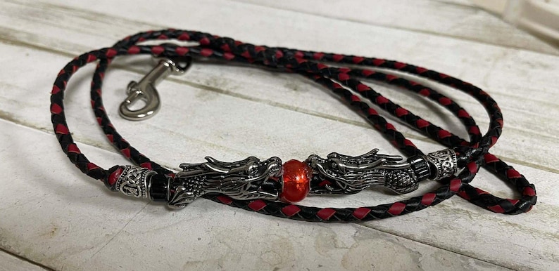 May include: Black and red braided leather leash with a silver-colored clasp. The leash features two silver dragon heads with a red bead in the center. The leash is designed for pets.