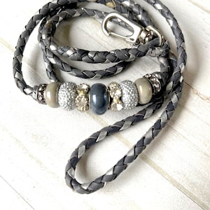 May include: Braided gray leather lanyard with silver accents and a silver clasp. The lanyard features decorative beads in shades of gray, silver, and clear crystals. A stylish accessory for keys or an ID badge.