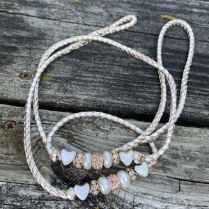 May include: Braided white and rose gold leash with heart-shaped charms, pearls, and rose gold accents. The leash is designed for a pet and is lying on a weathered wooden surface.