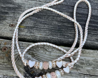 May include: Braided white and rose gold leash with heart-shaped charms, pearls, and rose gold accents. The leash is designed for a pet and is lying on a weathered wooden surface.