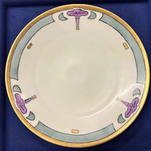 May include: A round ceramic plate with a cream-colored center and a decorative border. The rim features a gold-painted edge, light green curved segments, and three symmetrical purple floral motifs accented with small gold rectangular details.
