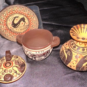 May include: A collection of four ceramic items including a round plate with a black deer illustration, a small two-handled bowl with brown interior, a circular lid featuring two bird figures, and a rounded vase decorated with black bird motifs.