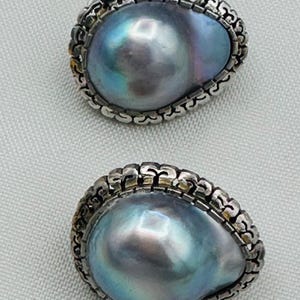 May include: A pair of earrings featuring large, iridescent, teardrop-shaped gray pearls. Each pearl is set within a decorative, scalloped silver-toned metal frame that features a repeating embossed pattern along the outer edge of the setting.