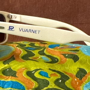 May include: Off-white sunglasses with dark lenses and a blue Vuarnet logo printed on the temple. The frame features a classic rectangular shape with silver-tone metal hinges connecting the arms to the front. The brand name is clearly visible.