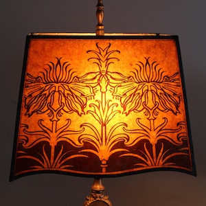 May include: A table lamp with a brown and orange lampshade featuring a stylized floral design. The lampshade is made of paper and has a black trim. The lamp base is made of metal and has a gold finish.