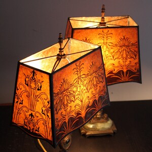May include: Two antique table lamps with brown and gold bases and matching shades. The shades are made of parchment paper with a floral design and are lit from within.