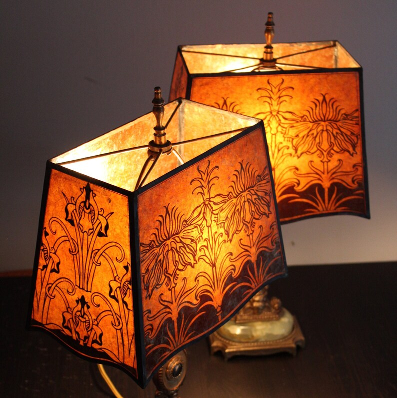May include: Two antique table lamps with brown and gold bases and matching shades. The shades are made of parchment paper with a floral design and are lit from within.