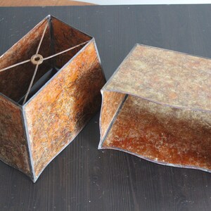 May include: Two brown lampshades with a textured surface. The lampshades are rectangular and have a black trim. One lampshade has a metal frame inside.