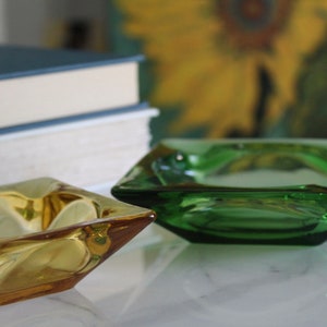 May include: Two square glass ashtrays, one is amber and the other is green. The ashtrays are sitting on a white surface.
