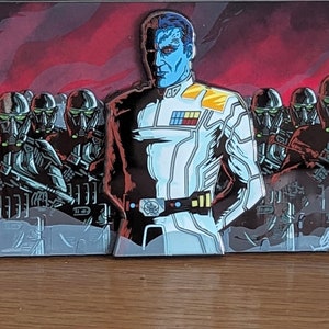 May include: A blue and red enamel pin featuring a character with a split face, one side blue and the other red. The character is wearing a white uniform with black accents and a gold belt. The character is standing in front of a group of black and red figures.