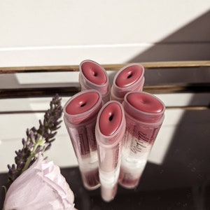 Puede incluir: Cinco tubos de brillo labial de color rosa rojizo est&aacute;n dispuestos en un patr&oacute;n circular. El brillo es visible a trav&eacute;s de los tubos transparentes. El producto est&aacute; etiquetado como "elderberry". Una ramita de lavanda est&aacute; en el fondo.