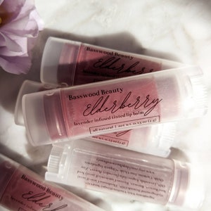 Puede incluir: Varios tubos de b&aacute;lsamo labial te&ntilde;ido con lavanda y sa&uacute;co de Basswood Beauty. Los tubos transparentes contienen un b&aacute;lsamo de color rosa-p&uacute;rpura. El texto "Elderberry" est&aacute; escrito en cursiva. Cada tubo contiene 4 g de producto. Sobre una superficie blanca.