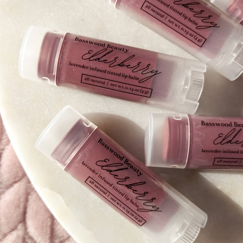 Puede incluir: Se muestran varios b&aacute;lsamos labiales te&ntilde;idos con infusi&oacute;n de lavanda. Los tubos transparentes tienen tapas blancas y contienen un b&aacute;lsamo de color rosa intenso. El texto "Basswood Beauty Elderberry" est&aacute; impreso en los tubos, junto con "lavender infused tinted lip balm" y "all natural | net wt. 4 g".