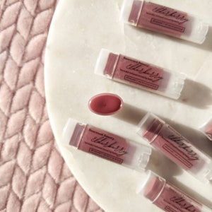 Puede incluir: Vista a&eacute;rea de varios b&aacute;lsamos labiales te&ntilde;idos con infusi&oacute;n de sa&uacute;co en tubos transparentes con tapas blancas. Los b&aacute;lsamos labiales est&aacute;n dispuestos sobre una superficie de m&aacute;rmol blanco. Un b&aacute;lsamo labial est&aacute; abierto, revelando el color malva. El texto "Basewood Beauty elderberry" es visible.