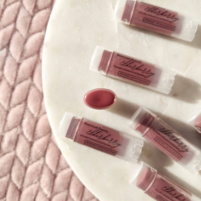 Puede incluir: Vista a&eacute;rea de varios b&aacute;lsamos labiales te&ntilde;idos con infusi&oacute;n de sa&uacute;co en tubos transparentes con tapas blancas. Los b&aacute;lsamos labiales est&aacute;n dispuestos sobre una superficie de m&aacute;rmol blanco. Un b&aacute;lsamo labial est&aacute; abierto, revelando el color malva. El texto "Basewood Beauty elderberry" es visible.
