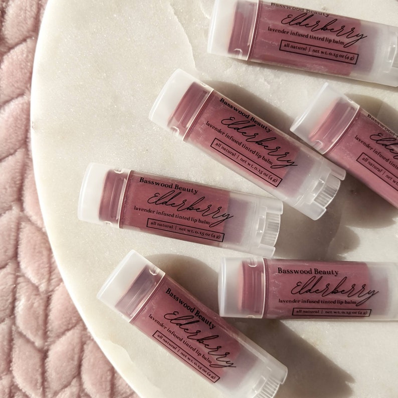 Puede incluir: Varios tubos de b&aacute;lsamo labial te&ntilde;ido con lavanda Elderberry de Basswood Beauty est&aacute;n dispuestos sobre una superficie de m&aacute;rmol blanco. Los tubos transparentes tienen tapas blancas y contienen un b&aacute;lsamo labial de color rosa intenso. El texto en los tubos dice "Elderberry" y "all natural | net wt. 4 g".