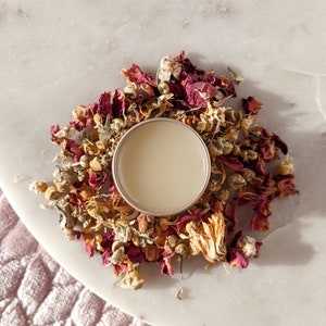 May include: A small, round, silver tin of lip balm surrounded by dried rose petals and other flowers.